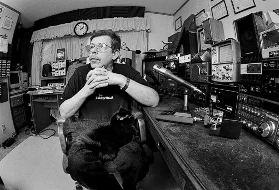 Black-and-white photograph of Art Bell seated in his radio studio with a black cat on his lap, a microphone beside him and radio equipment surrounding him